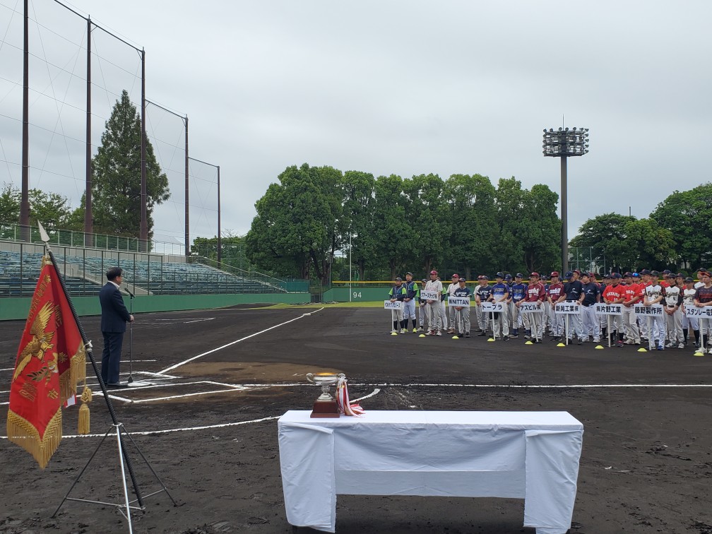 白いテーブルの上には優勝トロフィーが置かれ、マウンド付近にユニフォーム姿の選手たちが整列しており、ホームベース上でスーツ姿の市長がマイクでスピーチをしている写真