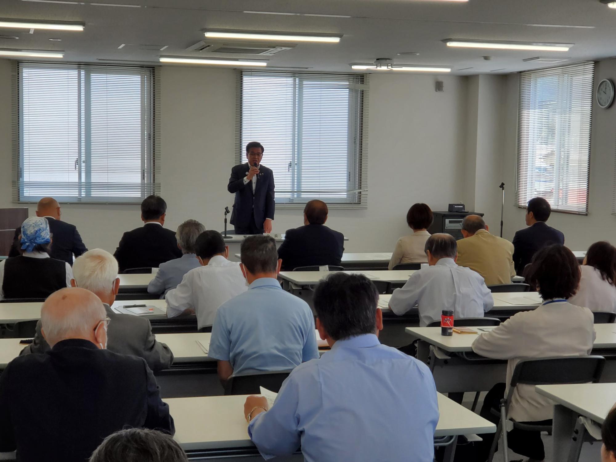 白と黒の机と椅子が並べられた会議室で、窓を背にしたスーツ姿の市長がマイクを持って聴衆に向かって立っており、老若男女の参加者が着席して話を聞いている様子を室内後方から写した写真