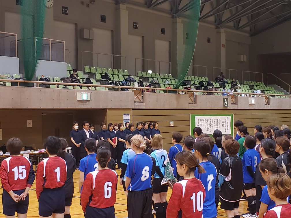 体育館で、スポーツユニフォームを着た複数の選手たちが、試合前の挨拶や説明を聞くために整列している写真