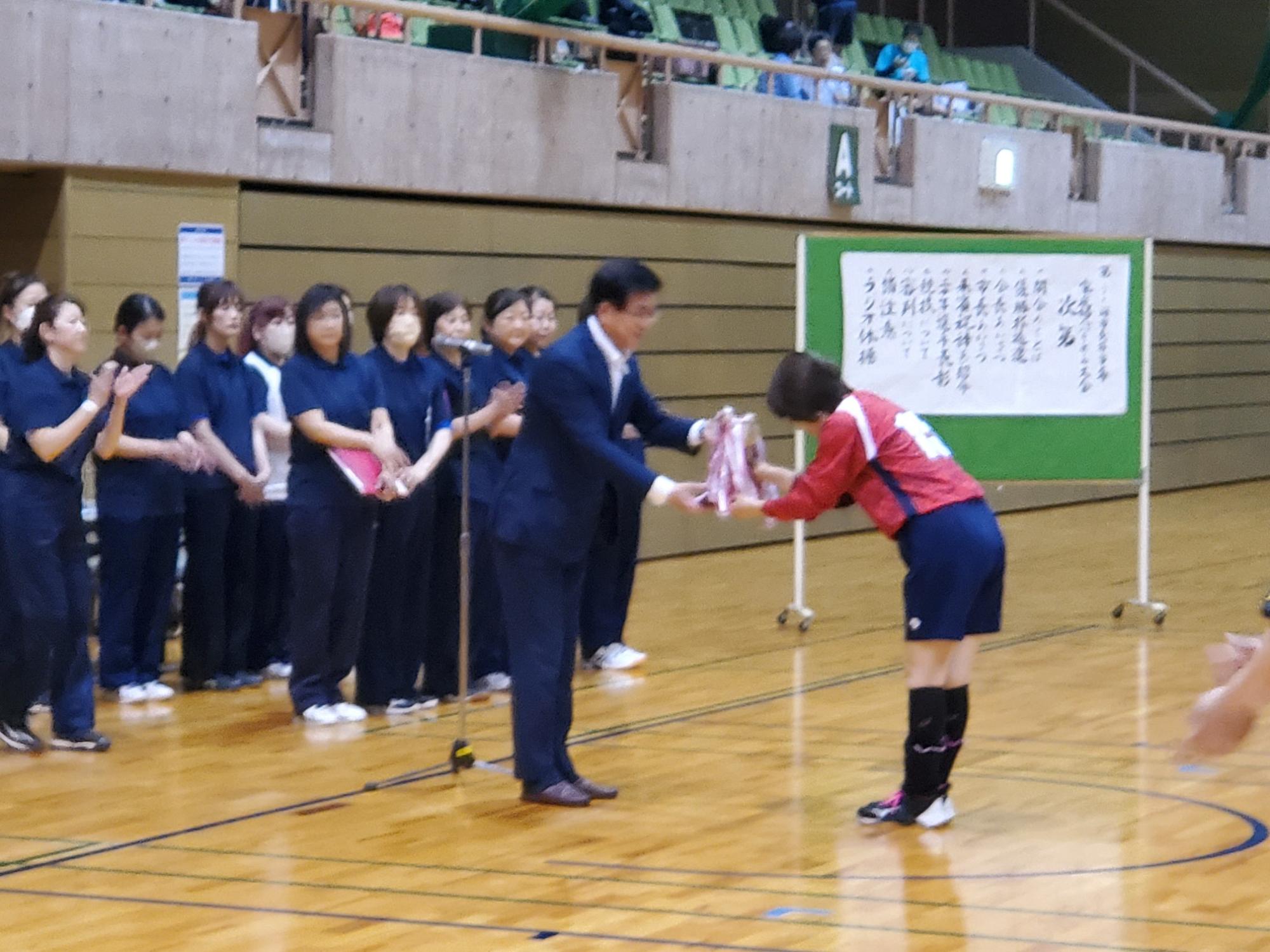 体育館の床の上で、女性選手がスーツ姿の市長からトロフィーを受け取っており、その周りを紺色のユニフォームを着た他の選手たちが囲んで拍手している写真