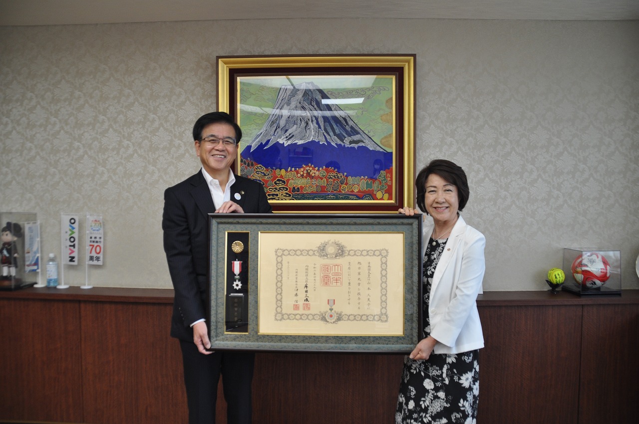 富士山の絵画を背景に、スーツ姿の市長と、白いジャケットを羽織った女性が、中央で勲章のついた大きな額縁を並んで持ち、笑顔で立っている写真