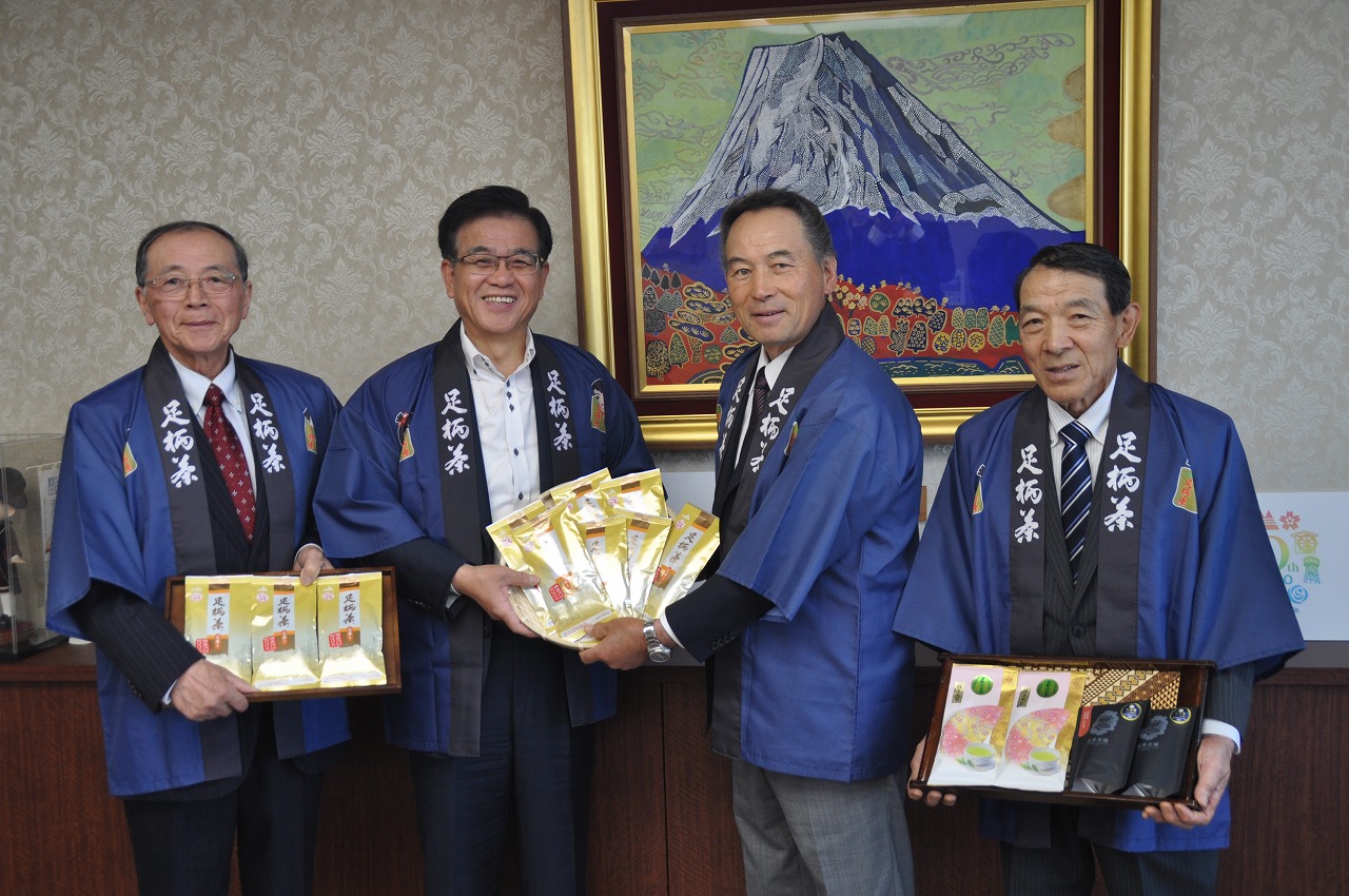 富士山の絵画が飾られた壁の前で、市長と三人の男性が紺色の法被を着て、お茶の入った箱やトレーを持って笑顔で立っている、贈呈式の写真