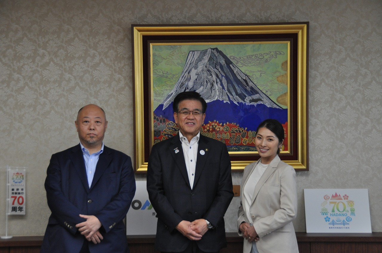 金色の額縁に収められた富士山の絵画が掛けられた淡い模様の壁の前で、スーツ姿の市長と男性と明るい色のジャケットを着た女性一人の三人が、にこやかに正面を向いて立って写真撮影をしている写真