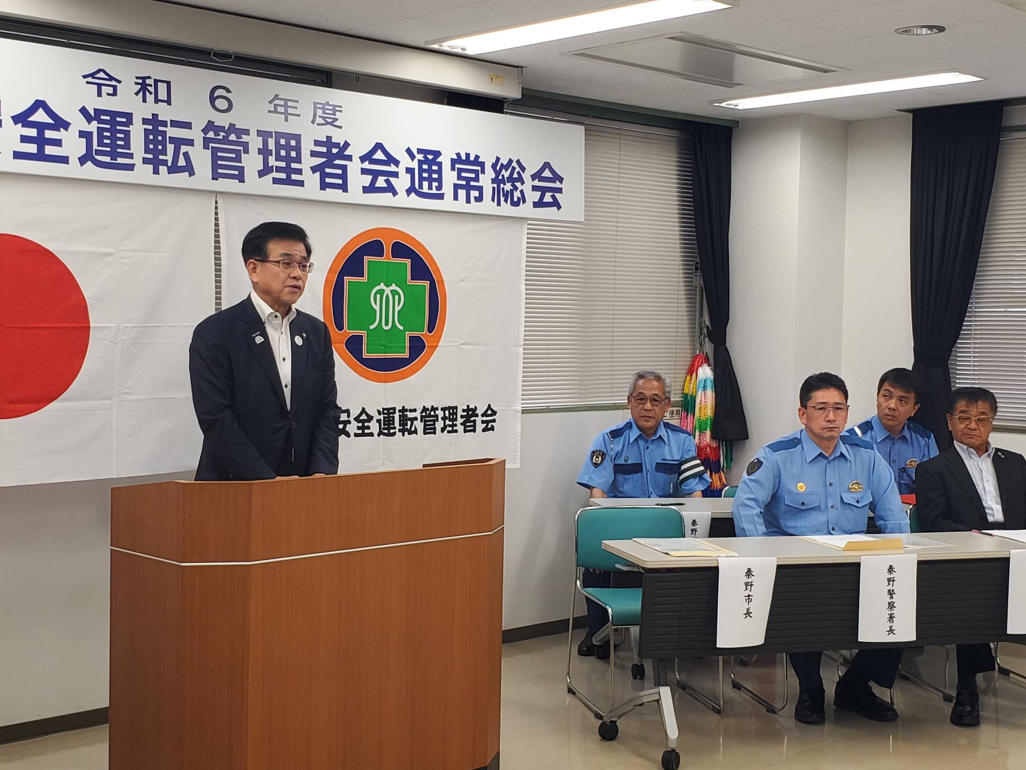 白地に「令和6年度 安全運転管理者会通常総会」と書かれた横断幕と日の丸を背景に、スーツ姿の市長が木製の演台に立って話しており、その右横の長机には制服姿の警察官を含む四人の男性が着席している会議の様子の写真