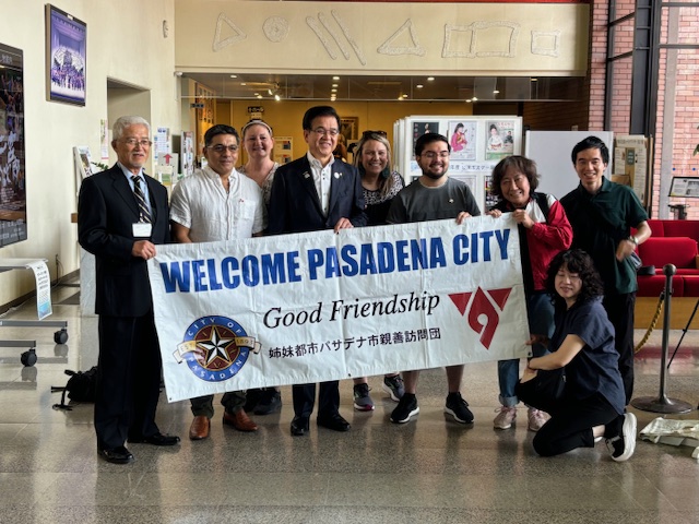 市役所のロビーのような場所で、スーツ姿の市長を含む日米の男女9人が、「WELCOME PASADENA CITY Good Friendship」と書かれた横断幕を持って写っている記念写真