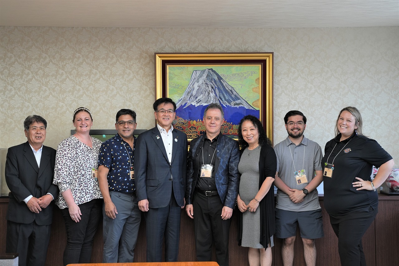 富士山の絵画が飾られた室内で、スーツ姿の市長と、カジュアルな服装の外国人が複数人並んで写っている記念写真