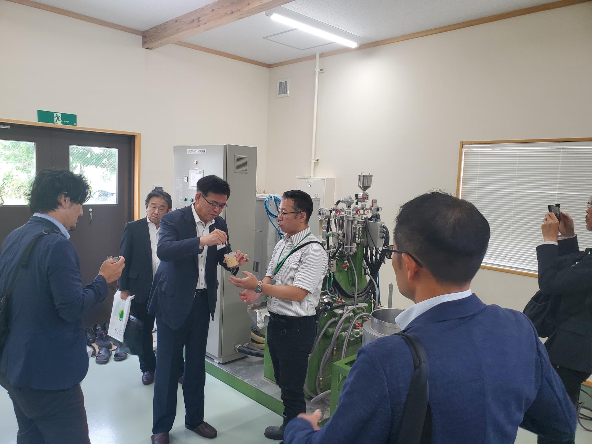 明るい室内に設置された機械の前で、スーツ姿の市長が、説明員から試食品らしきものを受け取っている見学風景の写真