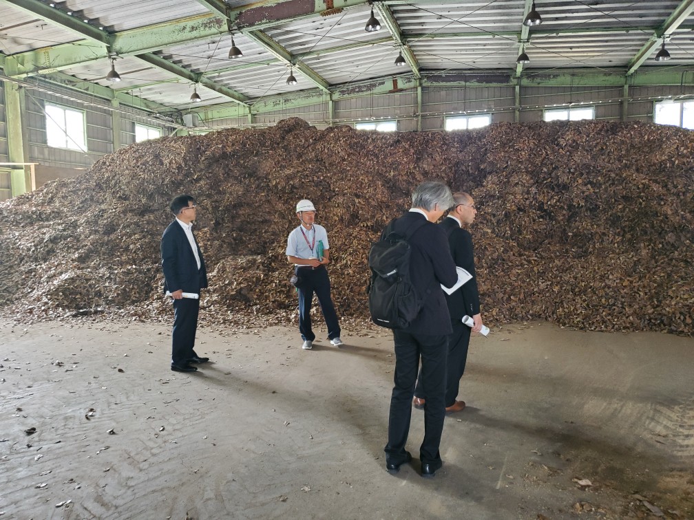 鉄骨の屋根と窓が設置された広い倉庫の中で、たくさんの落ち葉のようなものが山積みになっており、複数のスーツ姿の男性とヘルメットを被った男性が見学している写真