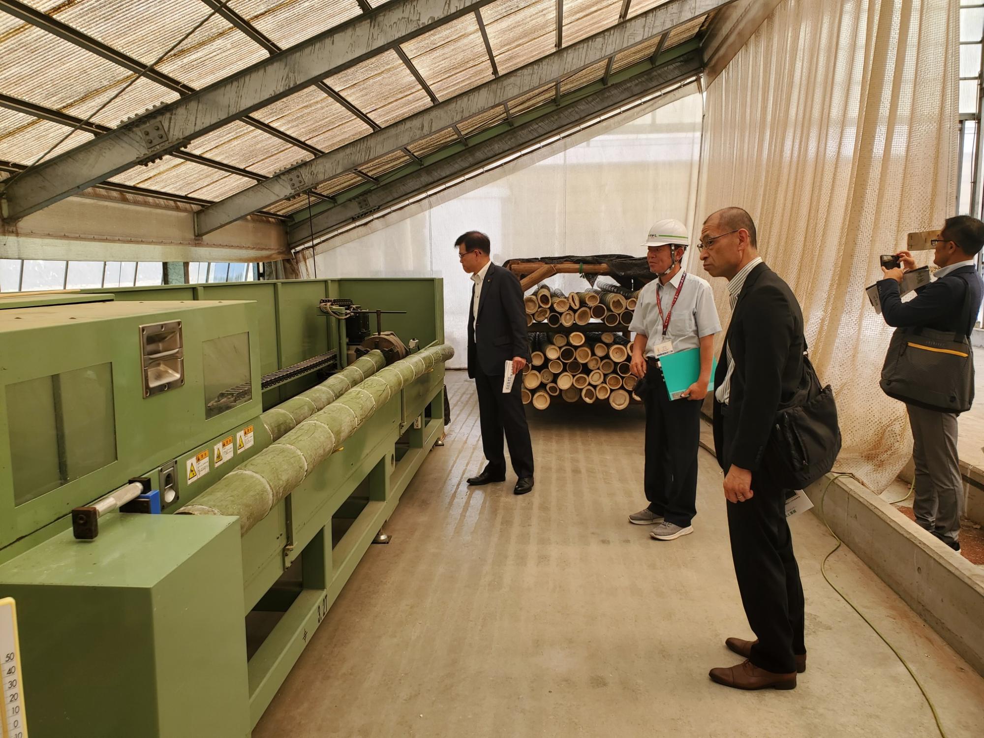 鉄骨造りの屋根の下、竹の加工機械の前で、スーツ姿の男性たちと作業服を着たヘルメット姿の男性が、機械や山積みになった竹を見学している写真