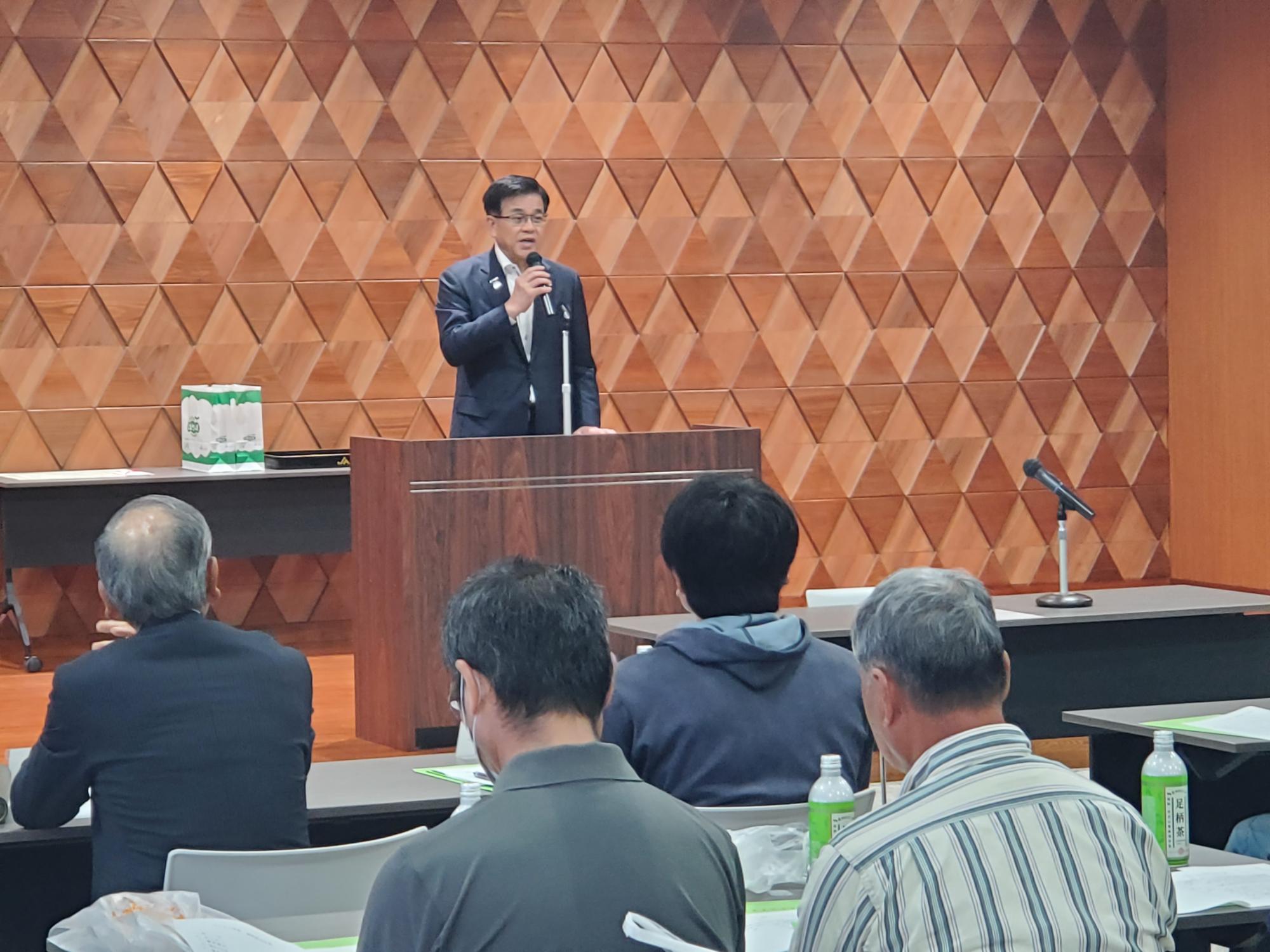 木目調の幾何学模様の壁を背景に、スーツ姿の市長が演台でマイクを持って話しており、複数の聴衆がその話を聞いている、会議の写真