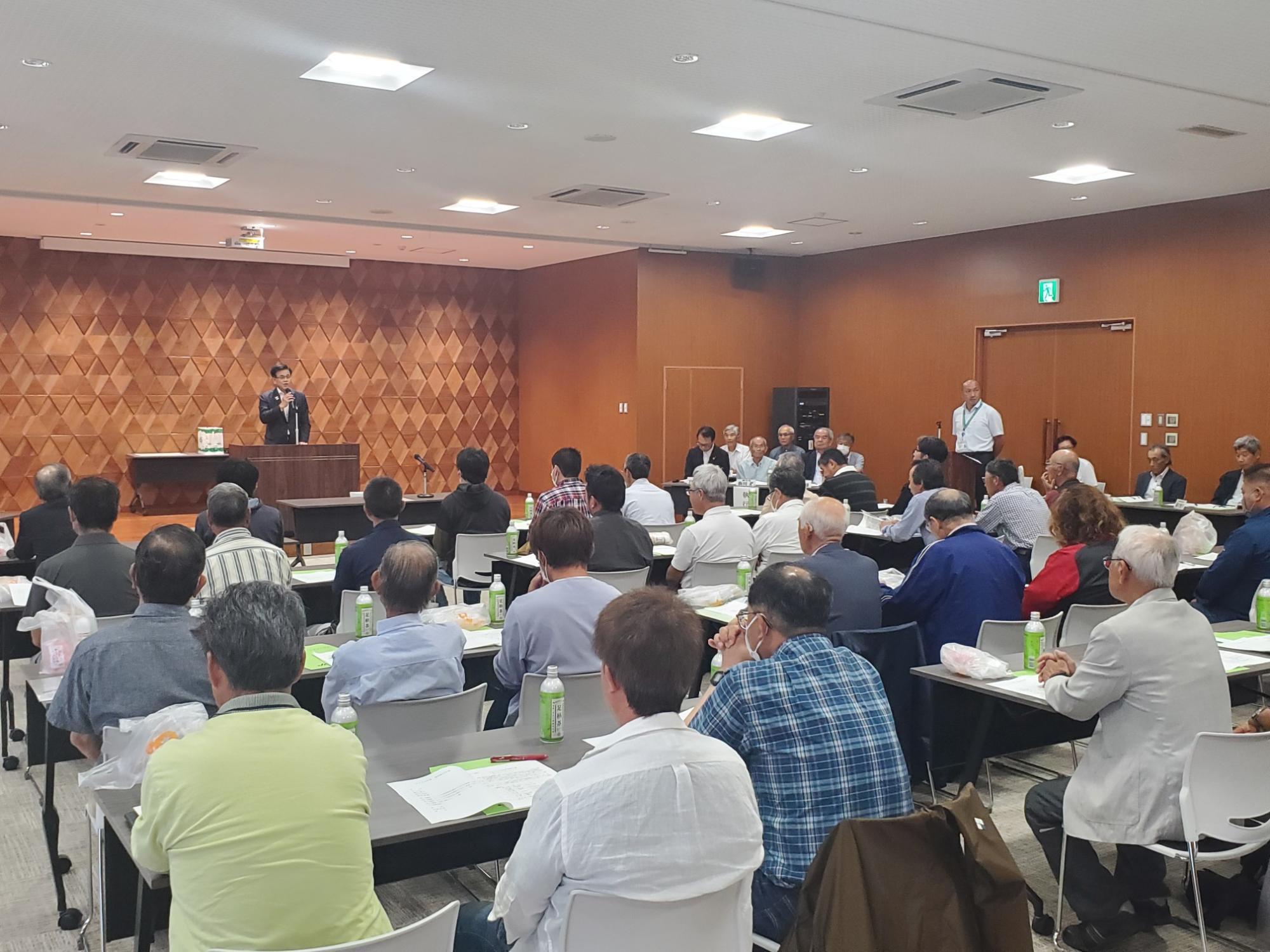 木目調の壁の部屋で、演台の前に立つ市長が、スクール形式に並べられた机に着席している大勢の聴衆に向けて話している、会議の写真