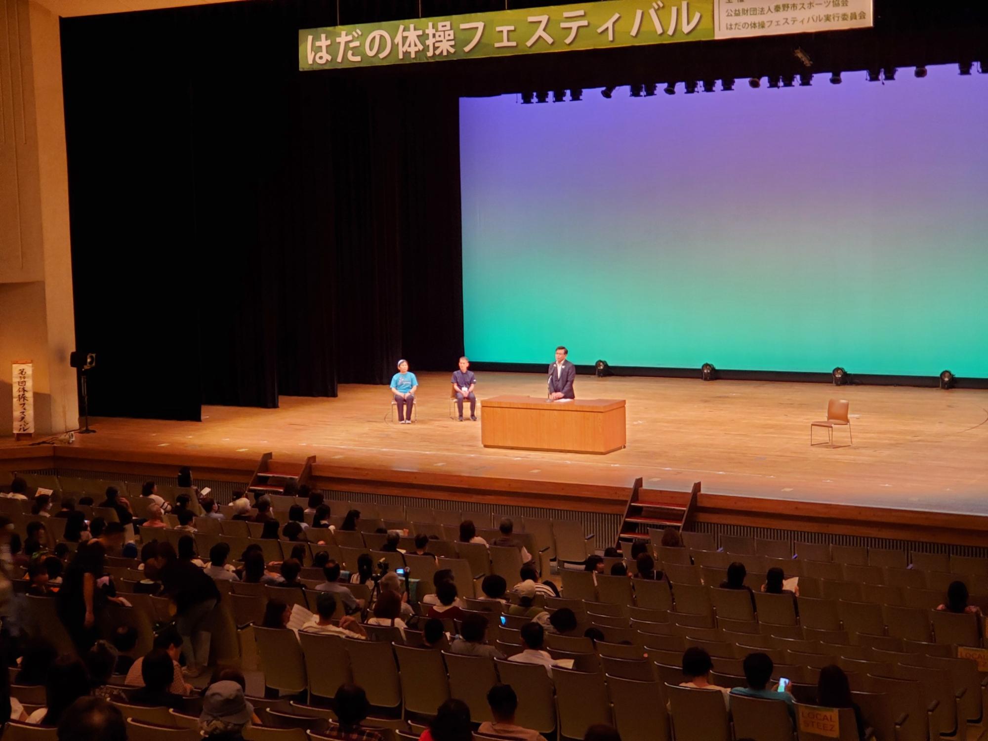 広いホールで、ステージ上に「はだの体操フェスティバル」の横断幕が掲げられ、たくさんの観客が見守る中、演台の周りで3人の大人が話している様子の写真