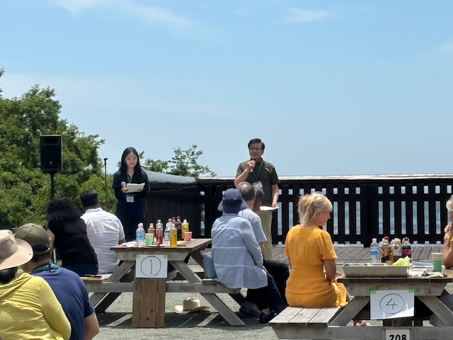 晴れた屋外で、木製のテーブルとベンチに座っている参加者を前に、市長と女性が、マイクを使って何かを説明しているイベント風景の写真