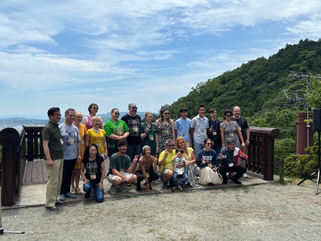 山の中腹の展望台のような場所で、パサデナ市民訪問団・青少年演奏団と地元の関係者が、街並みを背景に記念撮影をしている写真