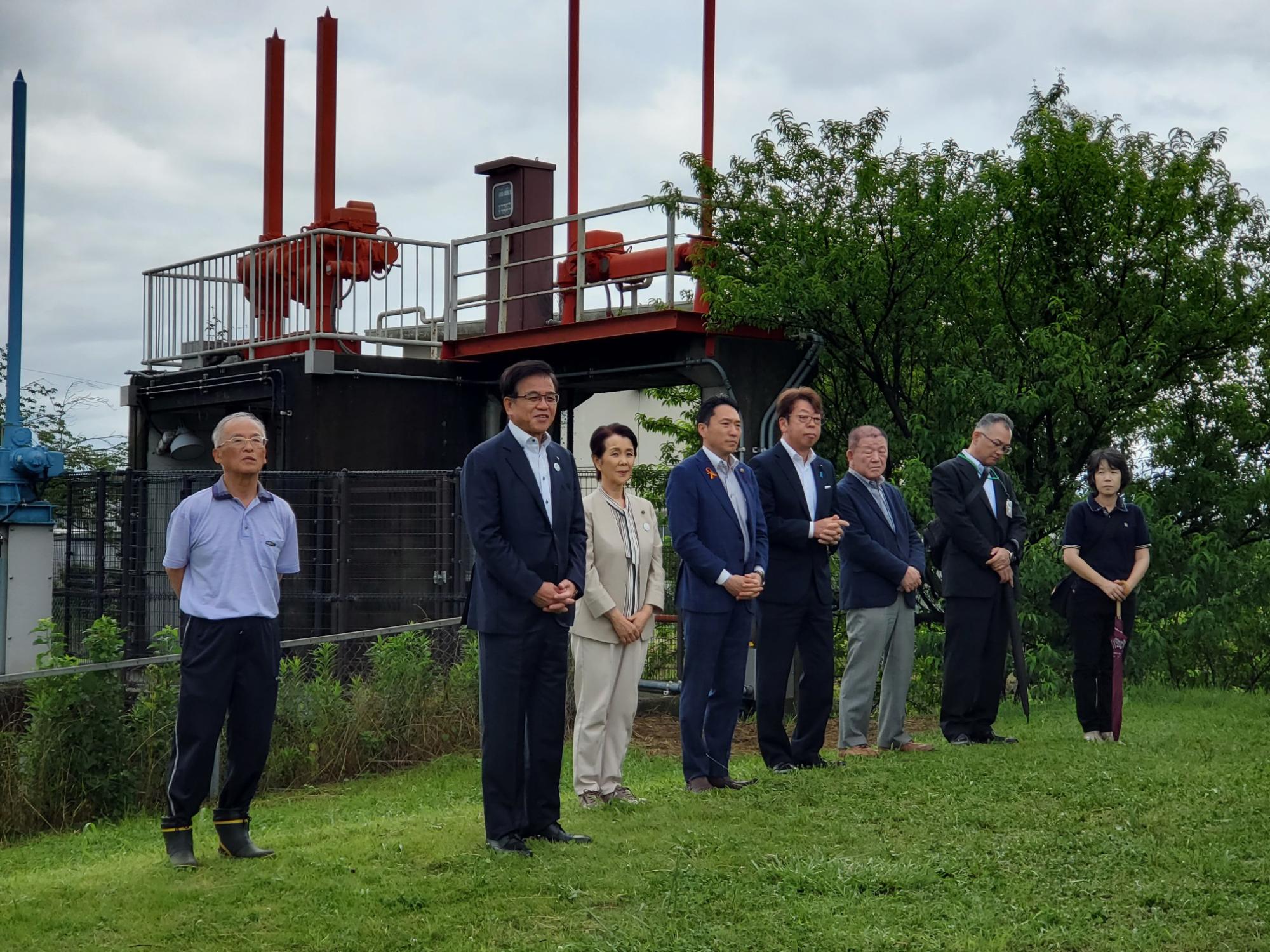 赤い配管と機械がある施設を背景にした芝生の上に立つスーツ姿の市長と4人の男性と女性、両脇に作業服の男性と黒い服の女性が並んでいる写真