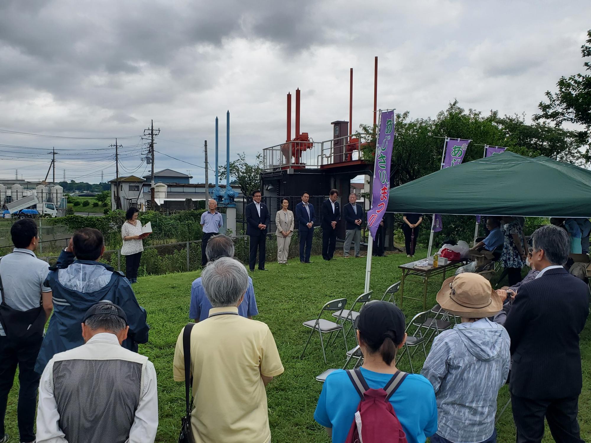 ポンプ設備らしきものが背景にあり、緑のテントの手前で、複数の参加者が立って挨拶を聞いている、式典の様子を写した写真