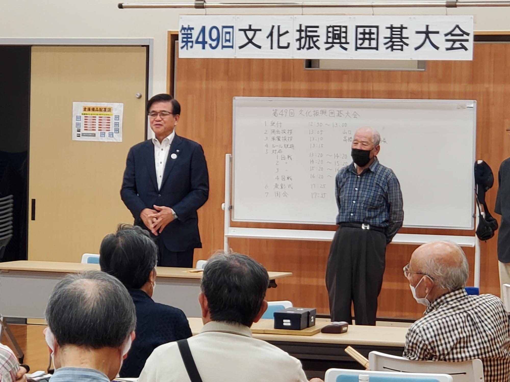 「第49回文化振興囲碁大会」の横断幕が掲げられた会場で、スーツ姿の市長が、マスクを着用した高齢の参加者たちに向かって挨拶をしている屋内写真