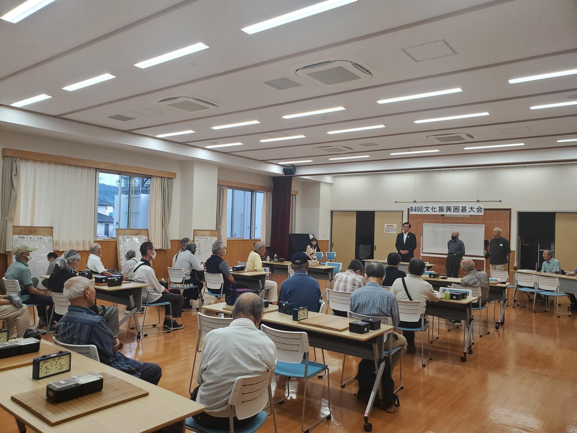 広い会場で、「第49回文化振興囲碁大会」の横断幕が掲げられており、大勢の高齢者が対局用の碁盤や時計が置かれたテーブルに向かい、挨拶をする人々の話を聞いている写真
