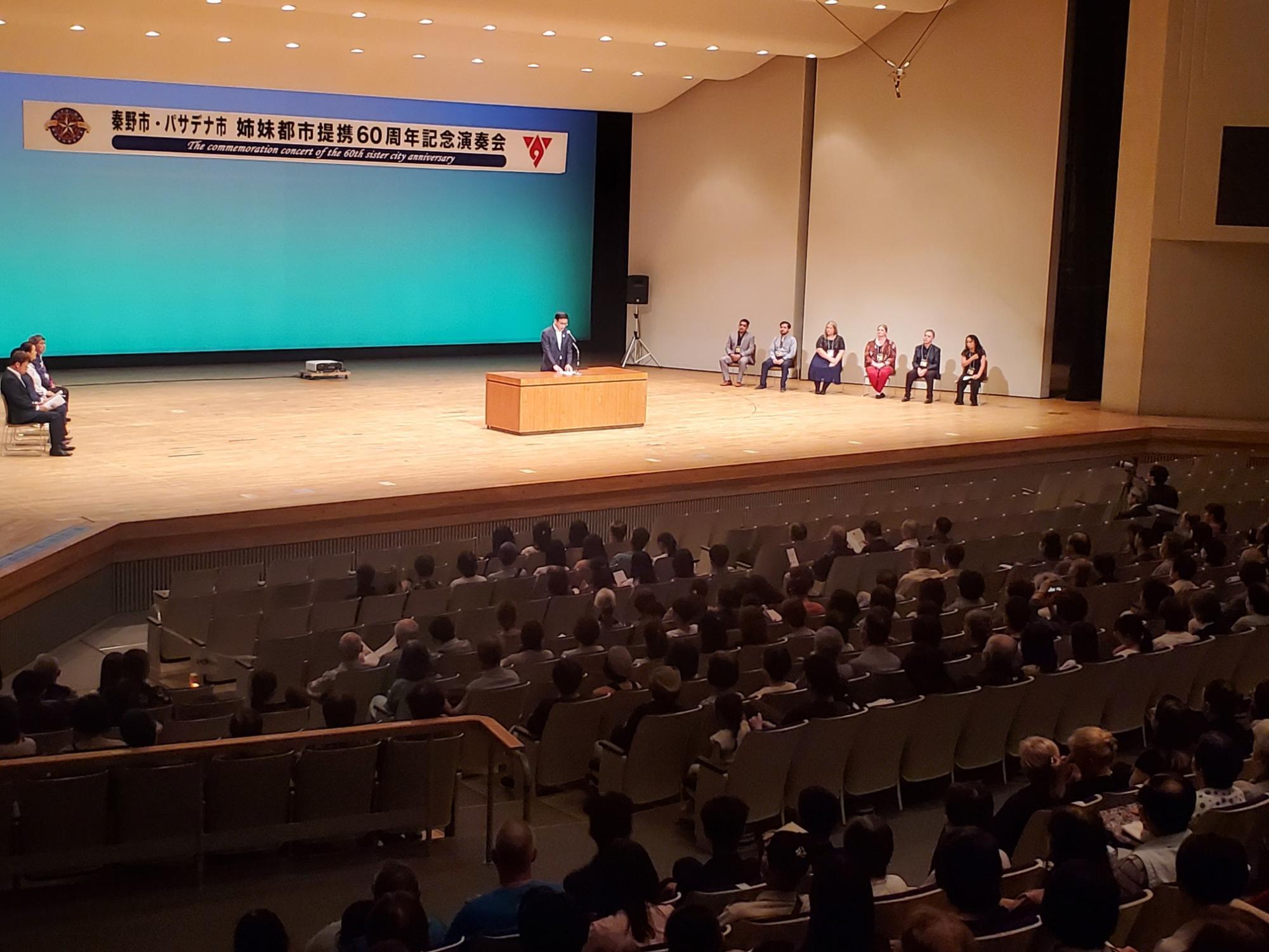 たくさんの観客が座るホールで、「秦野市・パサデナ市姉妹都市提携60周年記念演奏会」の看板が掲げられたステージ上に、演説台と複数の来賓が座っている写真