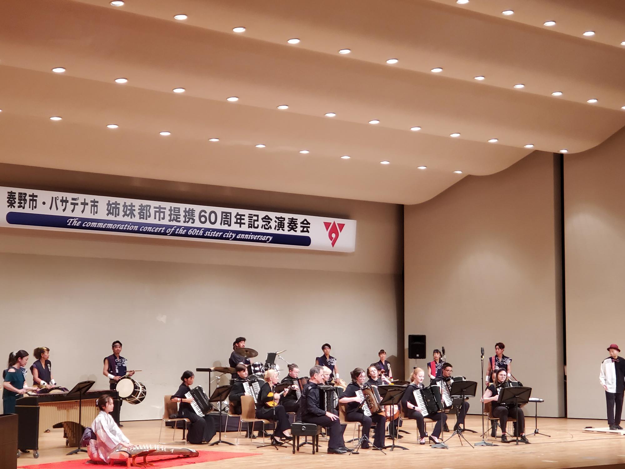 「秦野市・パサデナ市 姉妹都市提携60周年記念演奏会」の横断幕が掲げられたステージ上で、和楽器、ドラム、アコーディオン、弦楽器など、様々な楽器を演奏する人々が並んでいる写真