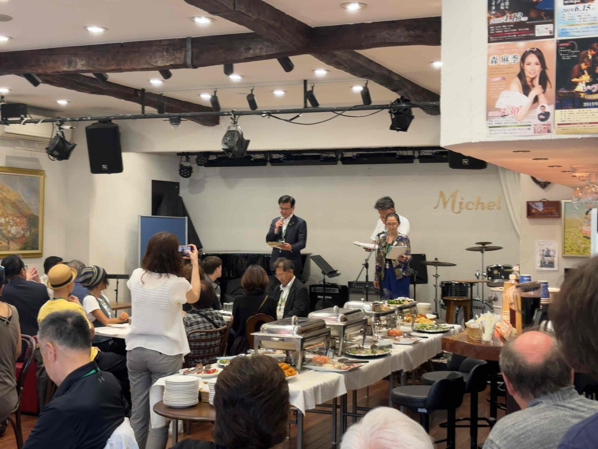 広い家屋内の会場で、ステージ上に立つ市長と二人の男女がマイクを使って挨拶をしており、手前のビュッフェ台を囲んで多くの客が着席している写真