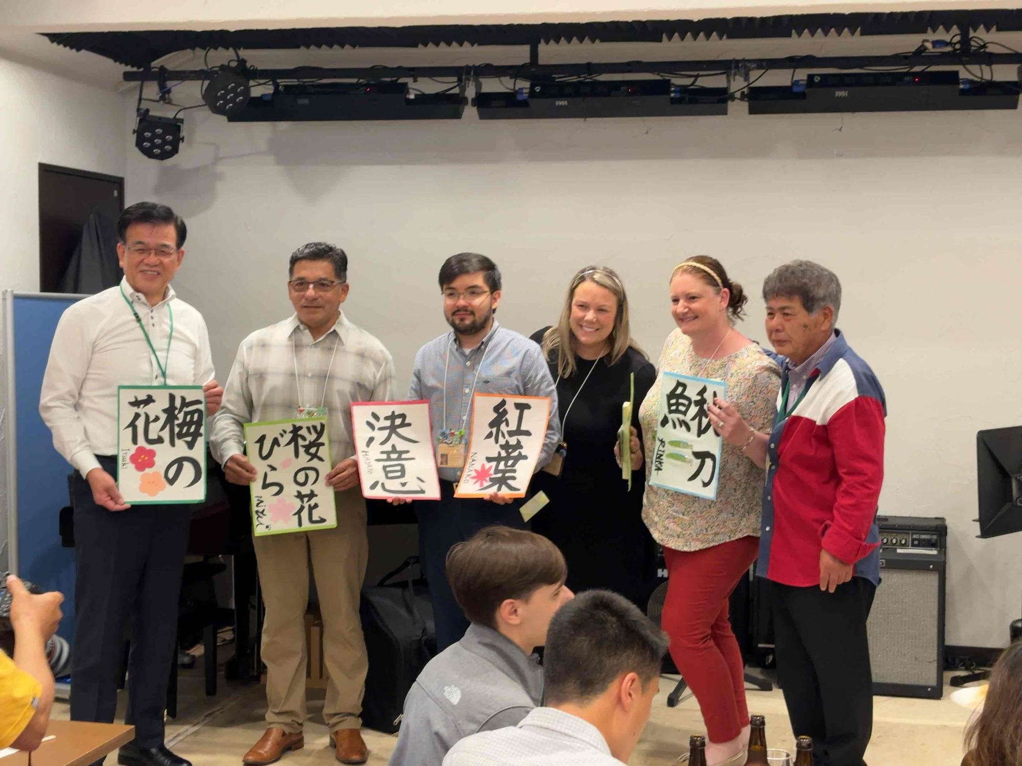 壇上の6人の男女が、それぞれの手に「梅の花」「桜の花びら」「決意」「紅葉」「秋刀魚」と習字で書かれた半紙を持って並んで写っている記念写真