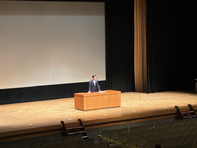 演台の後ろに大きなスクリーンと黒いカーテンが設置されたステージで、スーツ姿の市長がマイクを使って演説している屋内写真