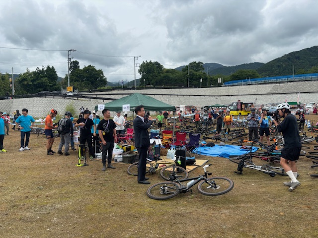 曇り空の下、広大な土の広場で、スーツ姿の市長がマイクを持って挨拶しているのを中心に、多くの参加者が集まり、周囲にはイベント用テントや大量の自転車が置かれている野外イベント会場の様子の写真