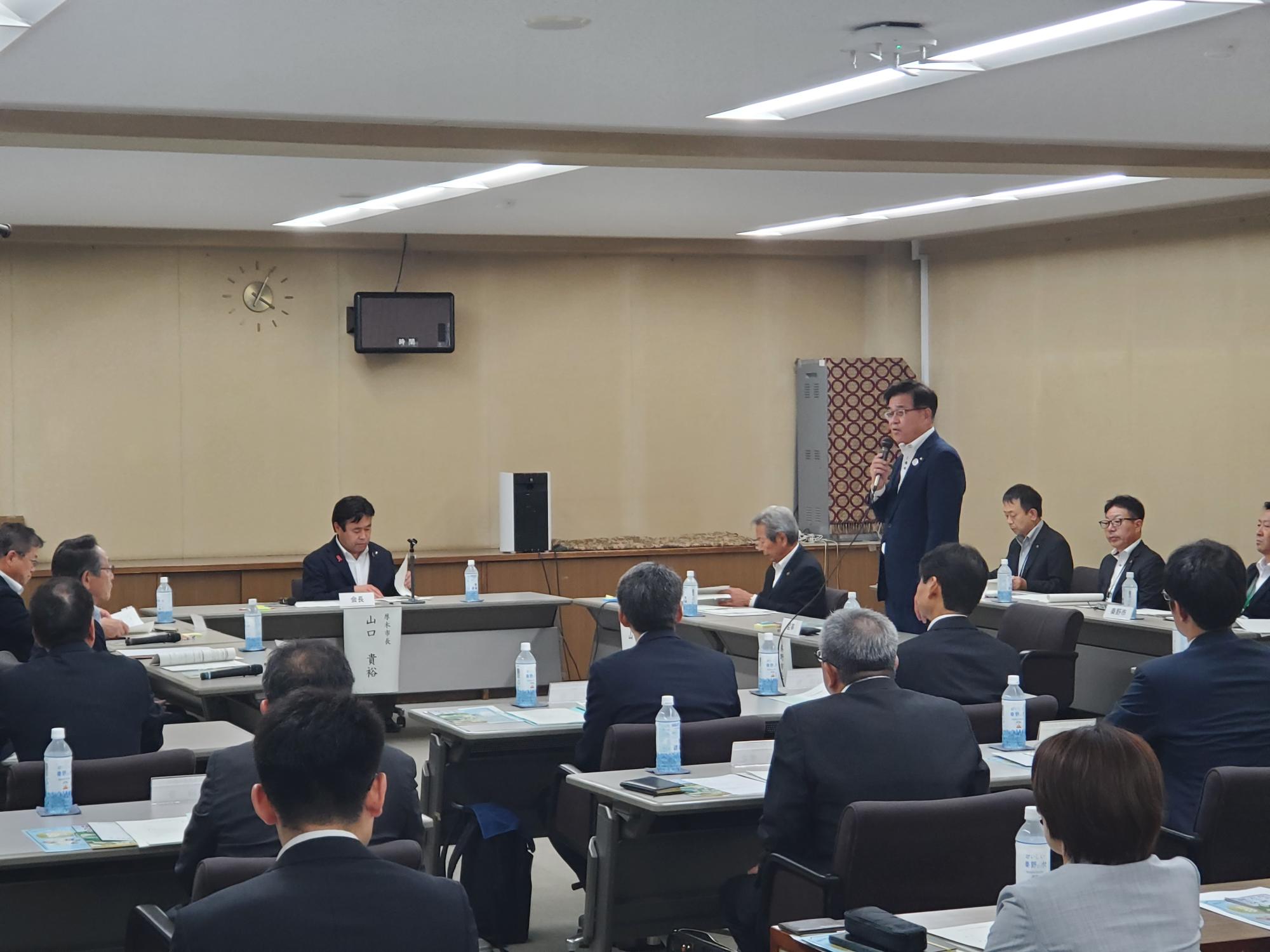 会議室で、複数のスーツ姿の出席者がテーブルに座り、市長がマイクを使って発言しており、会議資料や飲料水が整然と並べられた中で真剣に議論している様子