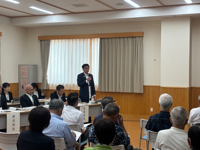 複数の椅子と机が並べられた部屋で、スーツ姿の市長がマイクを持って大勢の聴衆に向かって話をしている懇談会の写真