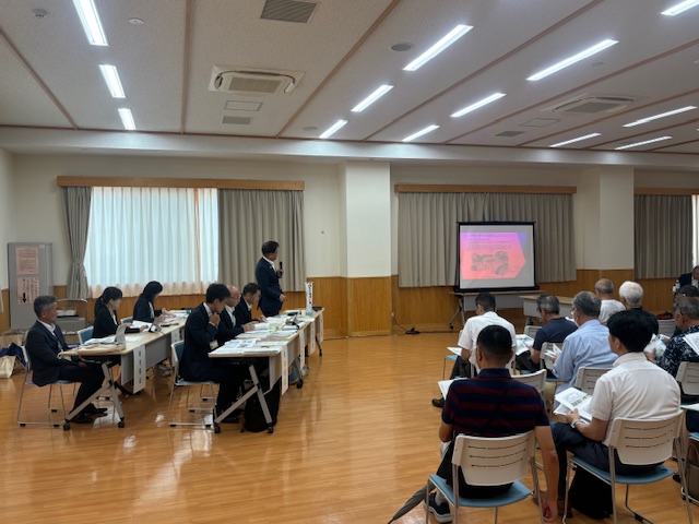 多数の参加者が並んだ椅子に座り、壇上の男性がマイクで話し、スクリーンに画像が投影されている、明るい室内での懇談会の様子