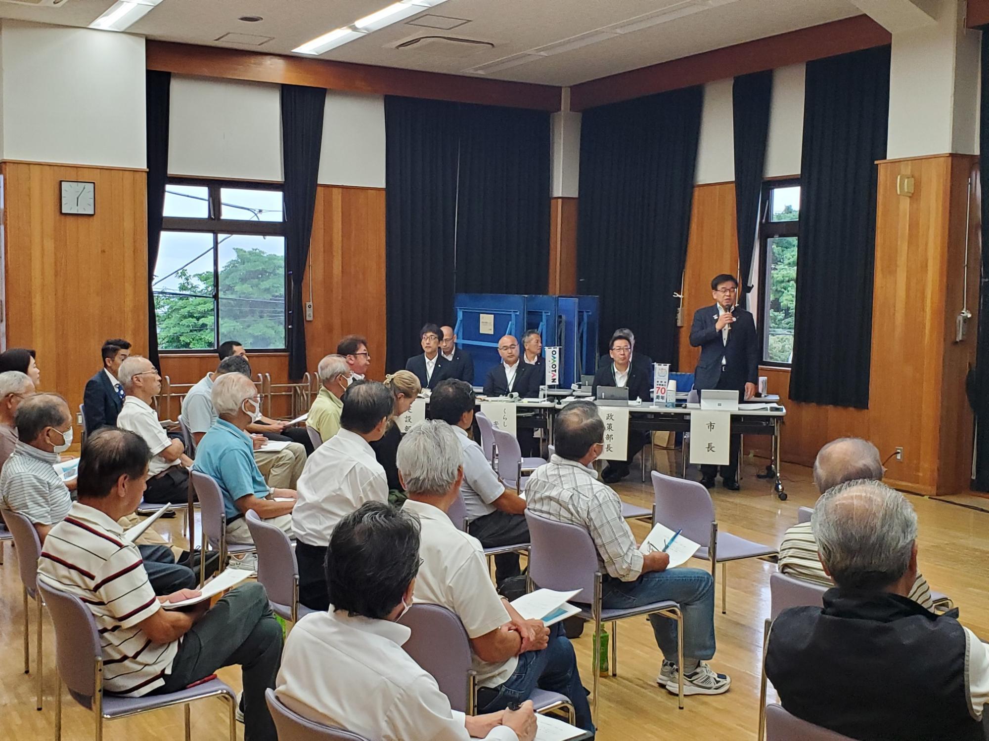 壇上ではマイクを持った市長がスピーチをしており、地域住民が集まる懇談会の様子の写真