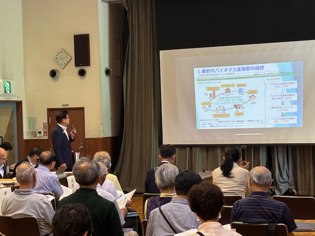 会議室で男性がマイクを持ってプロジェクターの画面を指し示し、参加者が資料を見ながら説明を聞いている懇談会の写真