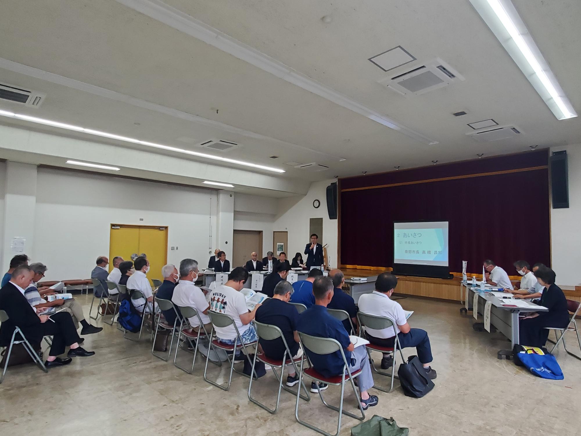 会場で、市長を含む行政関係者が前方に並び、地域住民が椅子に座って市政についての説明を聞いている様子の写真