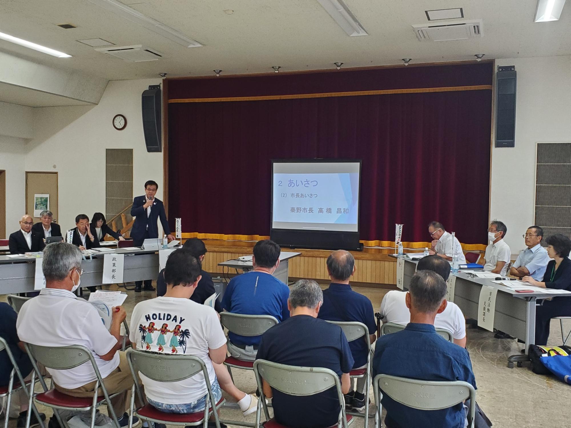 赤い舞台幕のある多目的ホールで、市長がマイクを持って挨拶をしており、スライドに「市長あいさつ」と表示され、地域住民と行政職員が向かい合って座って聞いている写真