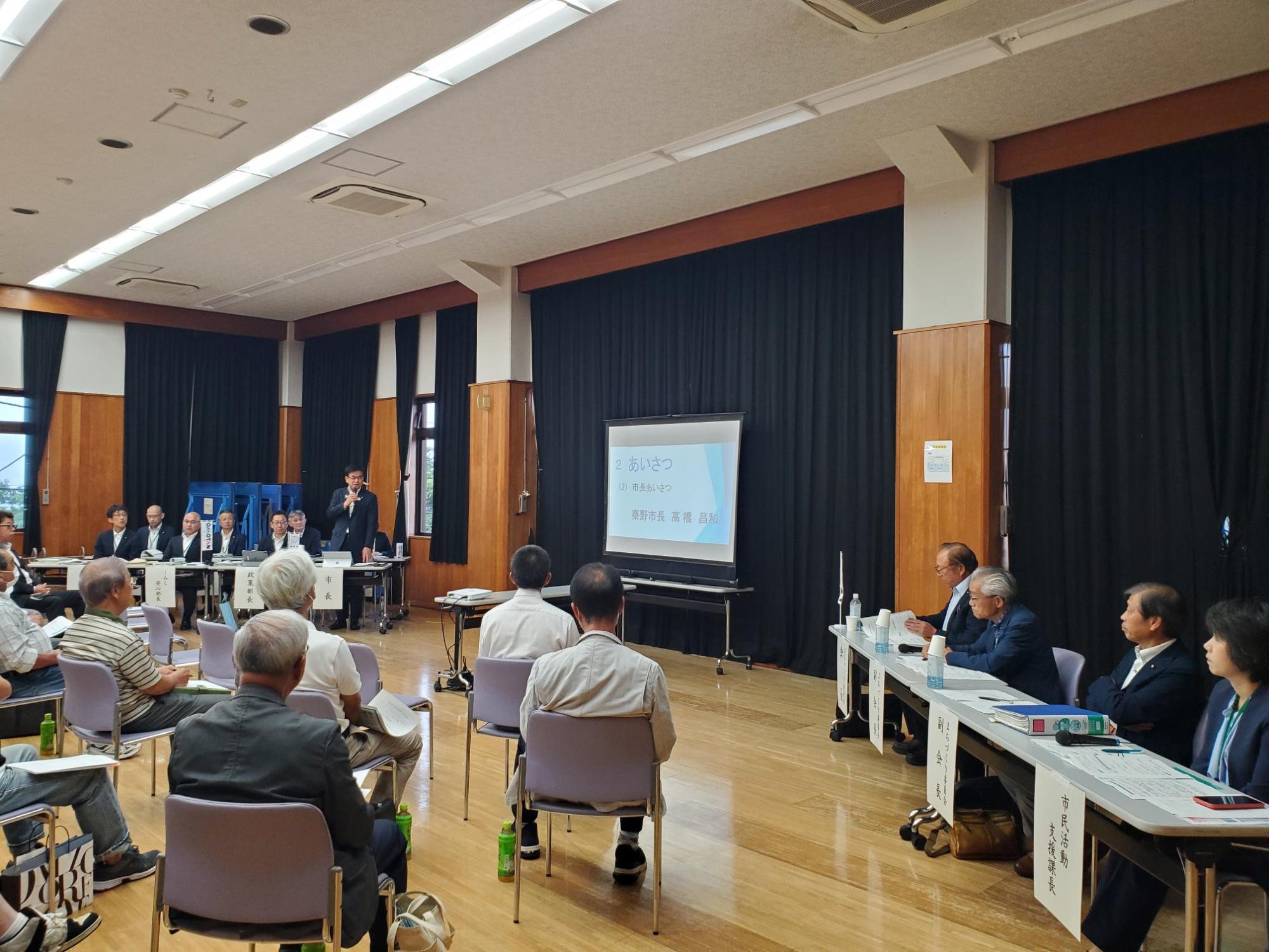 大勢の人が椅子に座り、壇上の市長がマイクを手に、スクリーンに投影された画面を見ながらスピーチをしている懇談会の写真