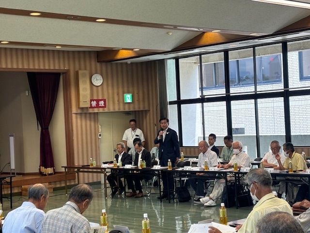 マイクを持った男性が発言し、複数の関係者が長テーブルに座り資料を見ながら意見交換している様子の写真