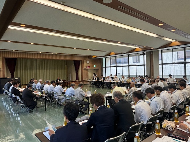 多数の関係者が円形に並べられたテーブルに座り、資料を読みながら議論や協議を行っている様子の写真