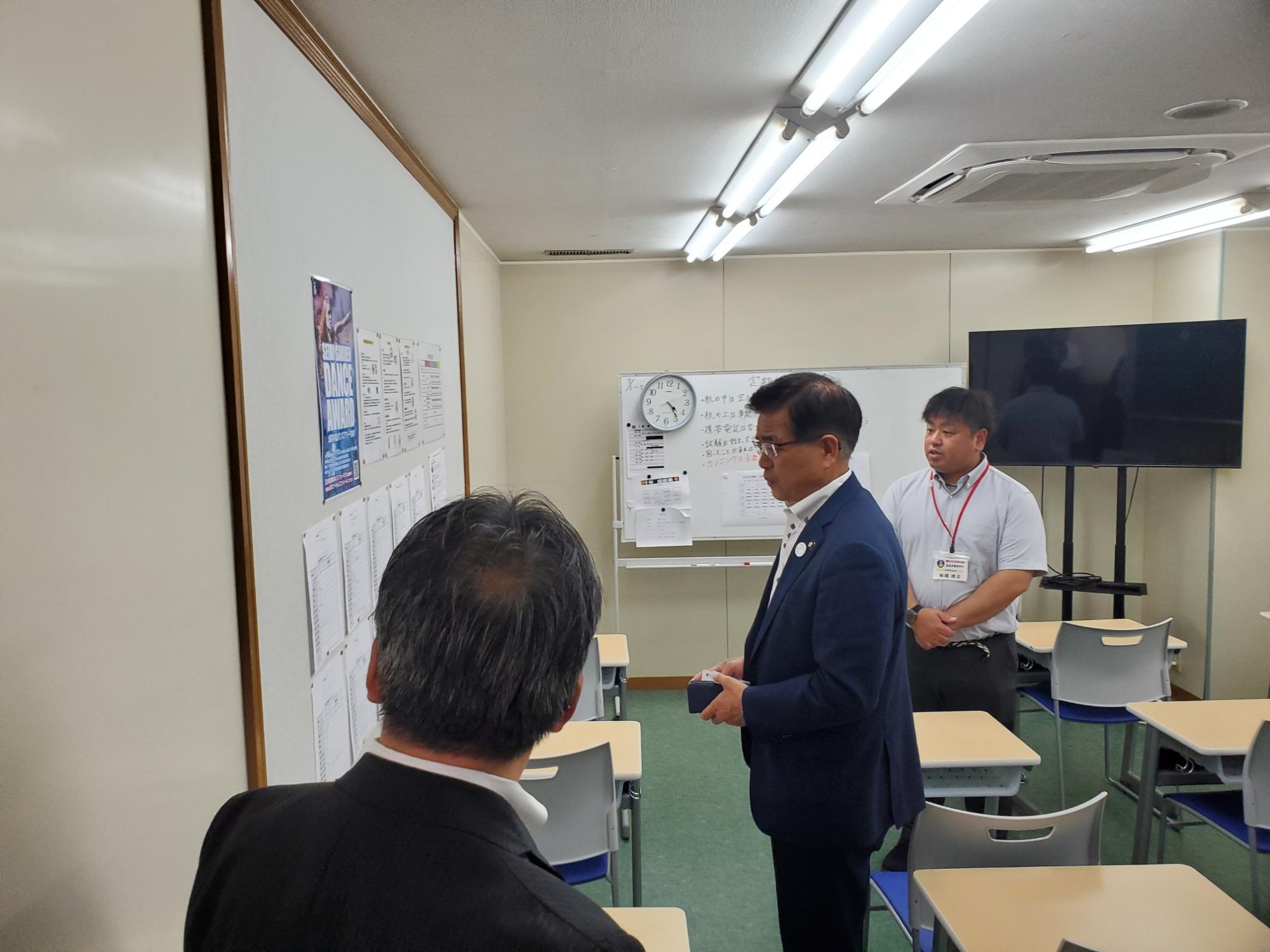 スーツ姿の3人の男性が壁に貼られた資料を確認している様子の写真