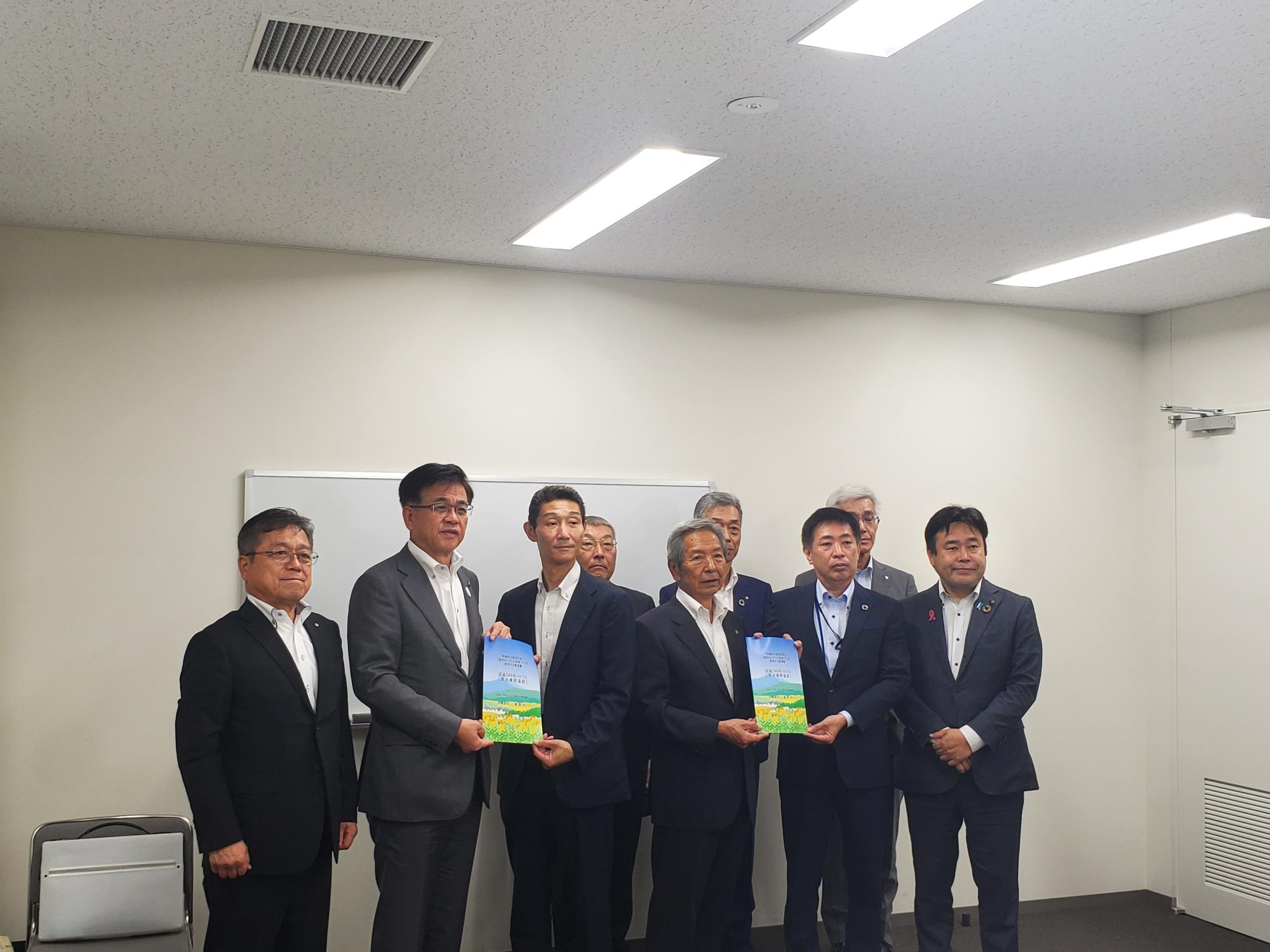 数名のスーツ姿の男性が並び、中央の二人が冊子を掲げてカメラに向かって立っている集合写真