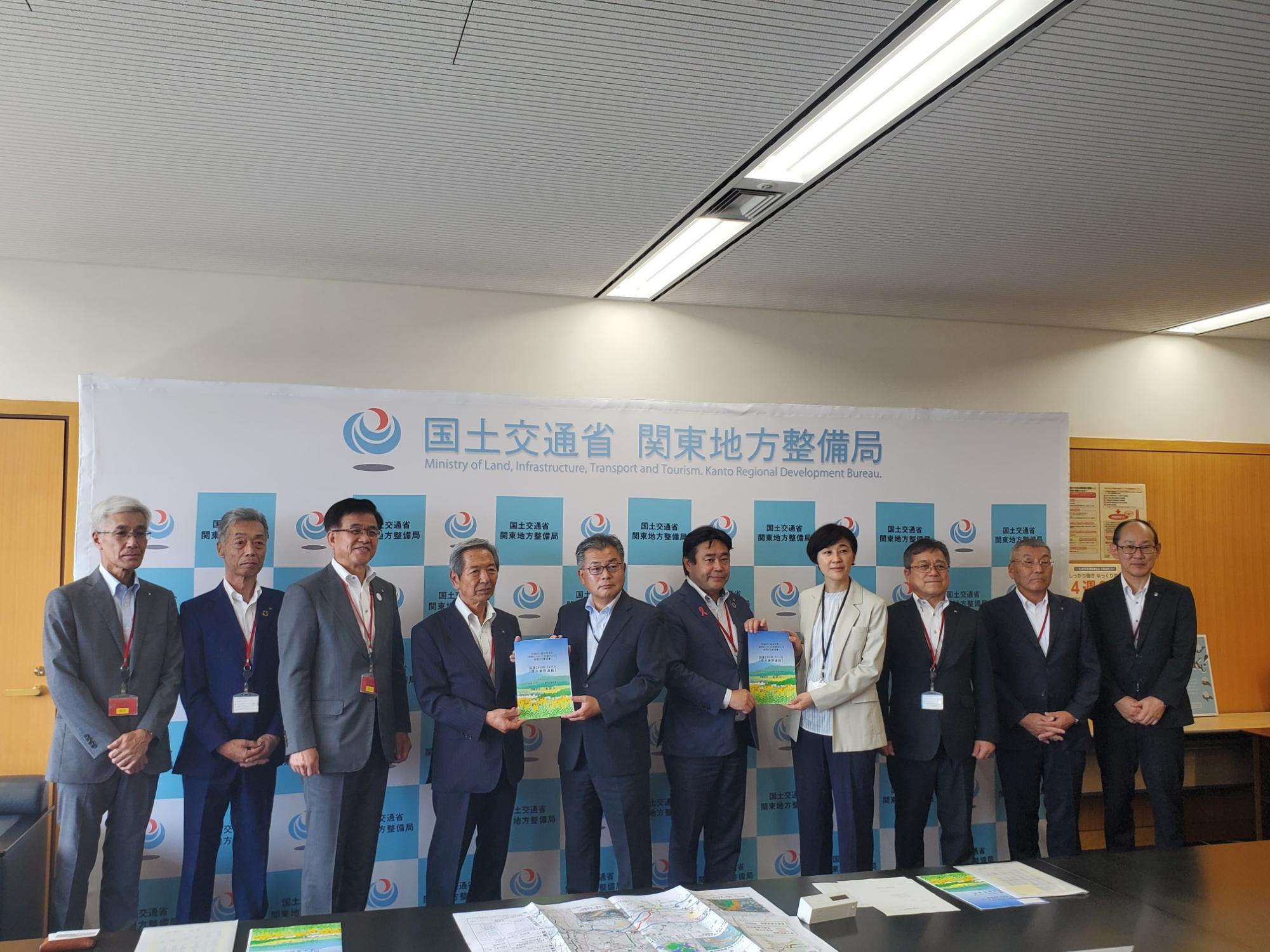 国土交通省関東地方整備局の看板を背景に、スーツ姿の関係者が並び、中央の2人が冊子を掲げている記念写真