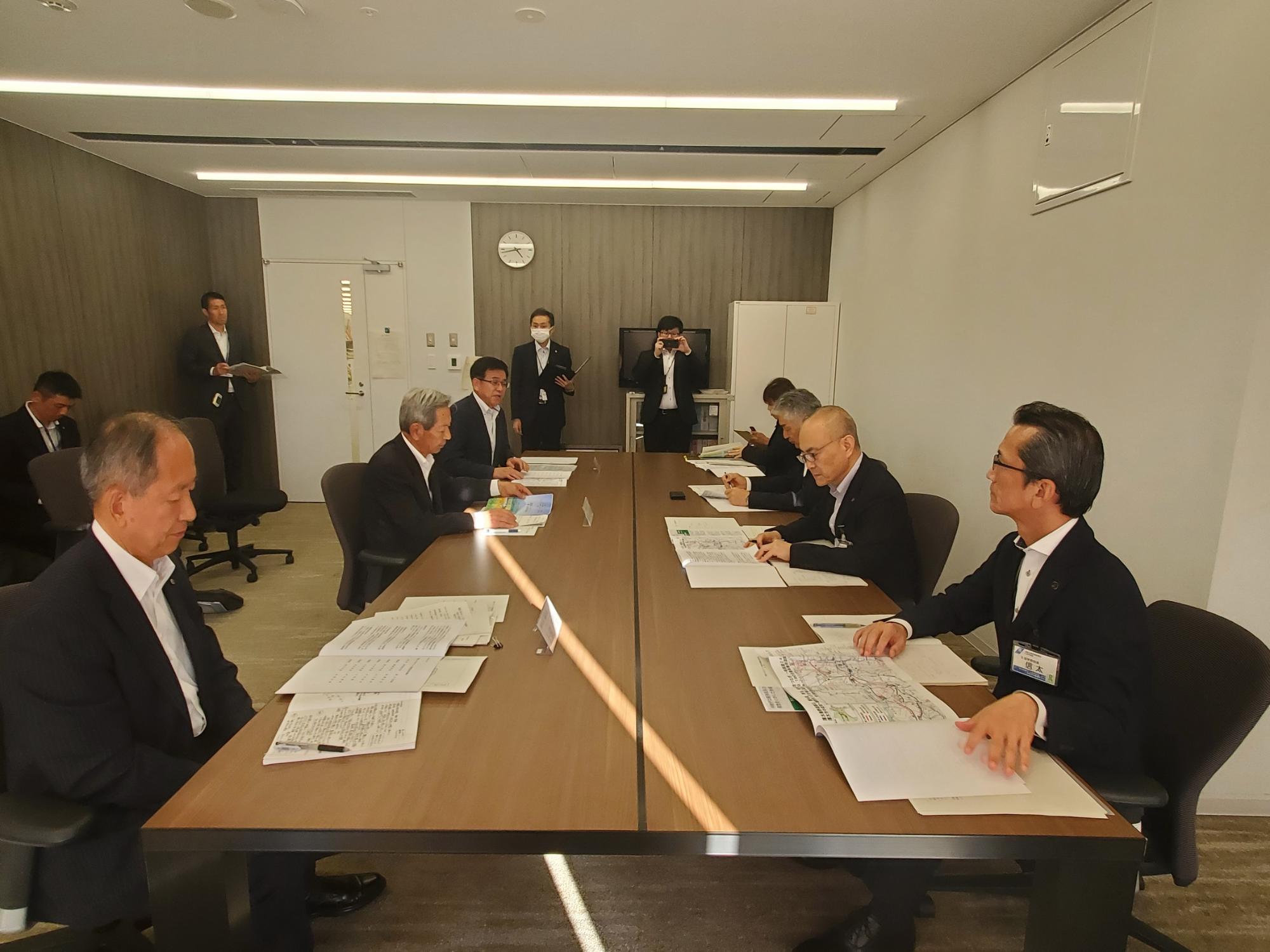 スーツ姿でテーブルを挟んで向かい合い、資料を前にして会議を行っている様子の写真