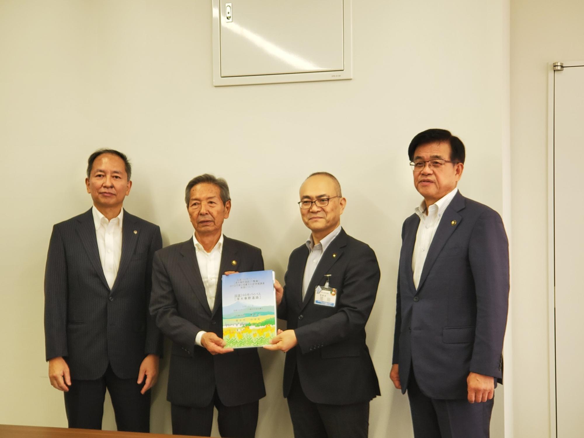 中央の2人がカラフルな冊子を一緒に持っており、スーツ姿の市長と男性3人が並んで記念撮影している写真