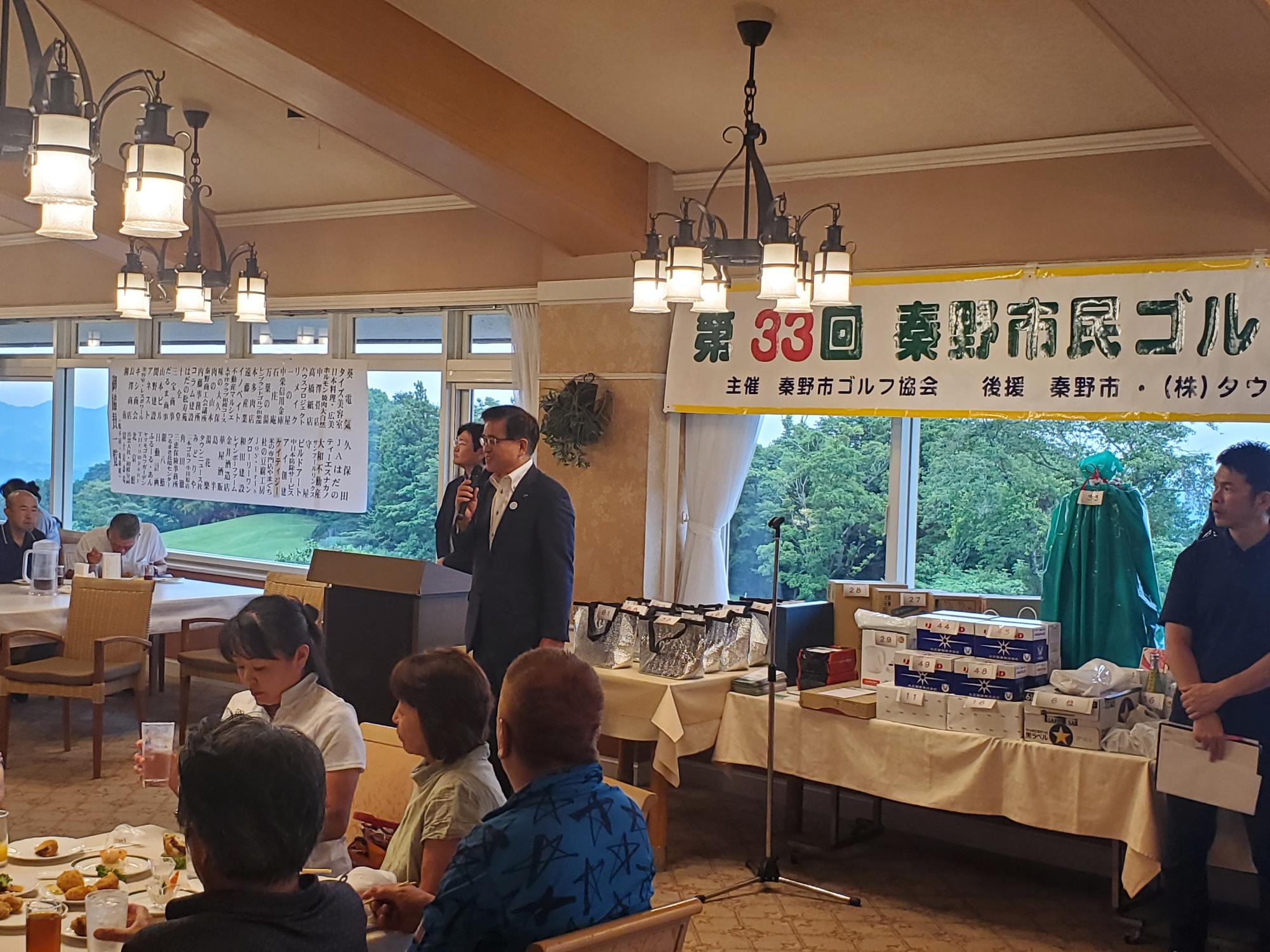 「第33回秦野市民ゴルフ大会表彰式」にてスーツ姿の市長がマイクを手に、食事をしている人々に向かって話をしており、その横に景品が並んでいる写真