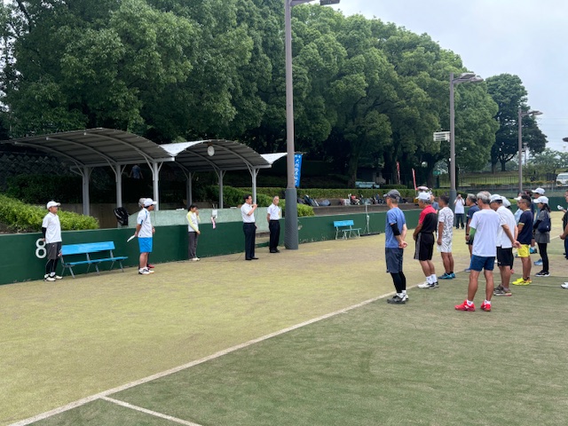 テニスコートを背景に、スポーツウェアを着た人々がグループに分かれて整列し、中央のスーツを着た男性が話している、開会式の写真