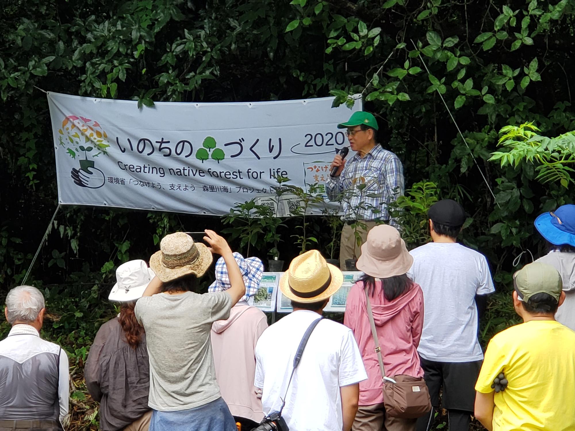 「いのちの森づくり」と書かれた横断幕が掲げられた森の中で、緑色の帽子をかぶった男性がマイクを使って聴衆に向かって話をしている様子の写真