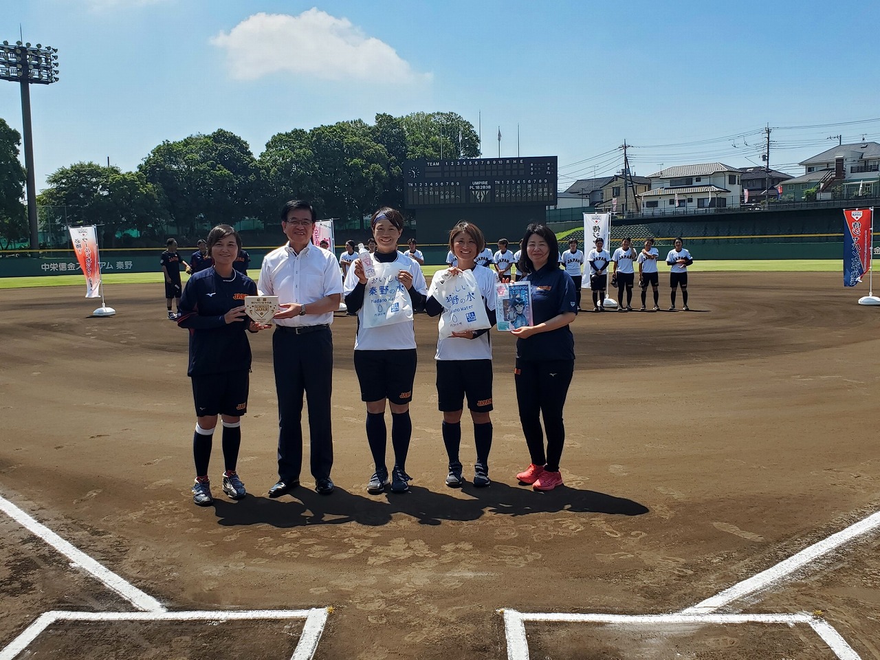 女子野球チームの選手2人と、市長と関係者が野球場のホームベース付近で賞品を手にして並び、背景にチームメンバーが整列している写真
