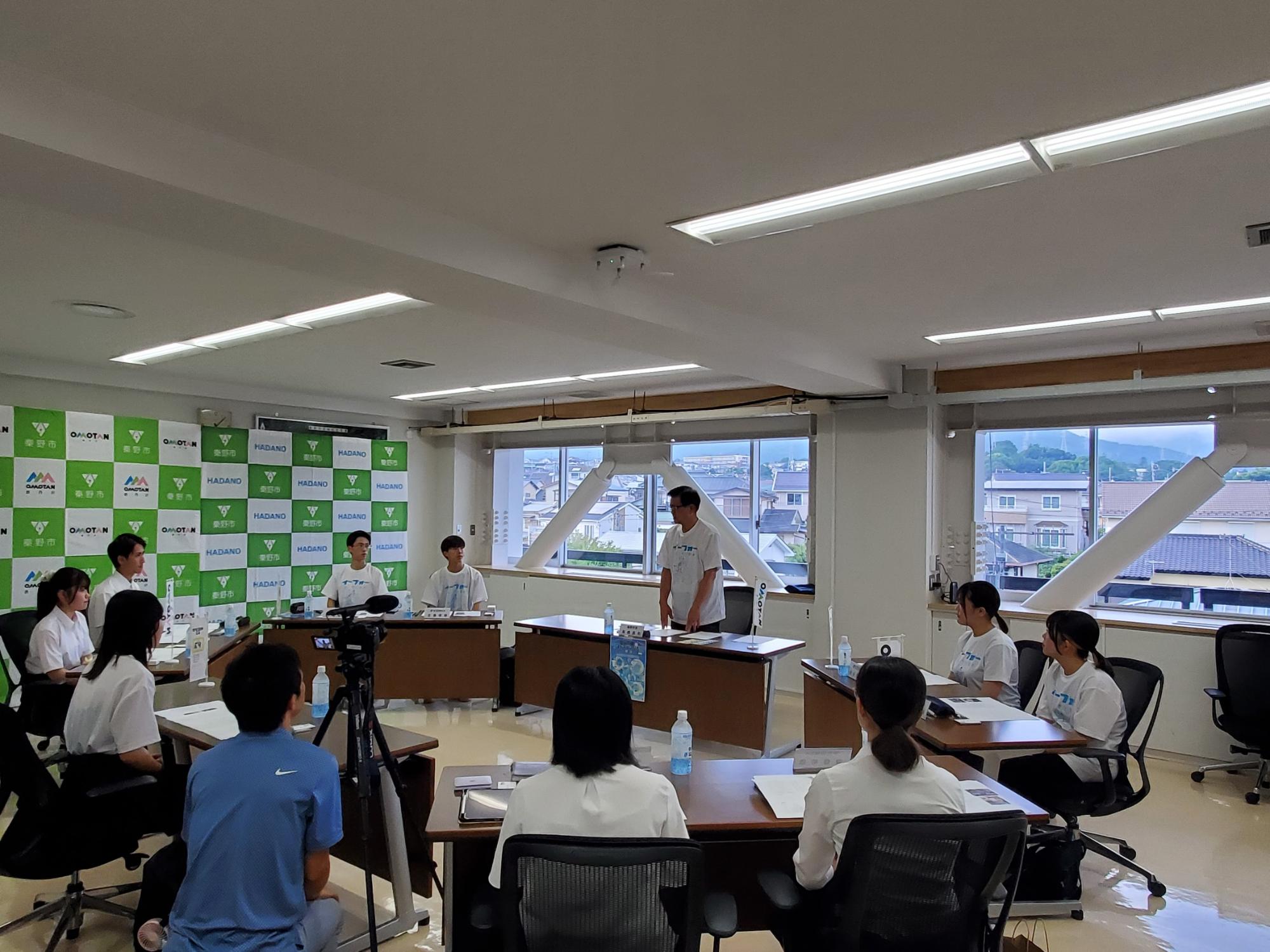 テーブルを挟んで向かい合うようにして座った複数の男女が、中央で話す市長の話を真剣に聞いている様子を捉えた写真