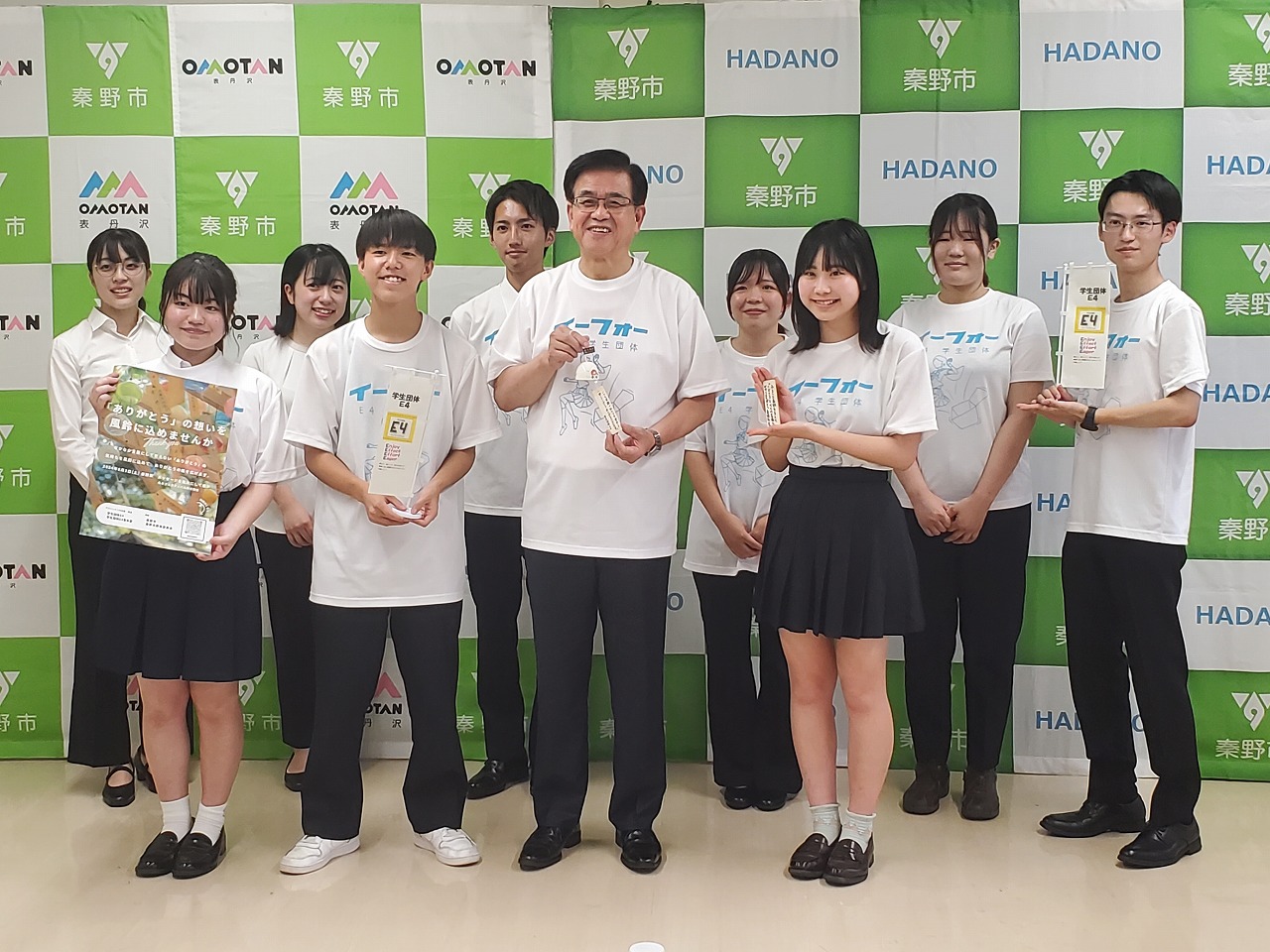 緑と白のチェック柄の秦野市のパネルの前で、表彰状のようなものを持ちながら記念撮影をしている様子の写真