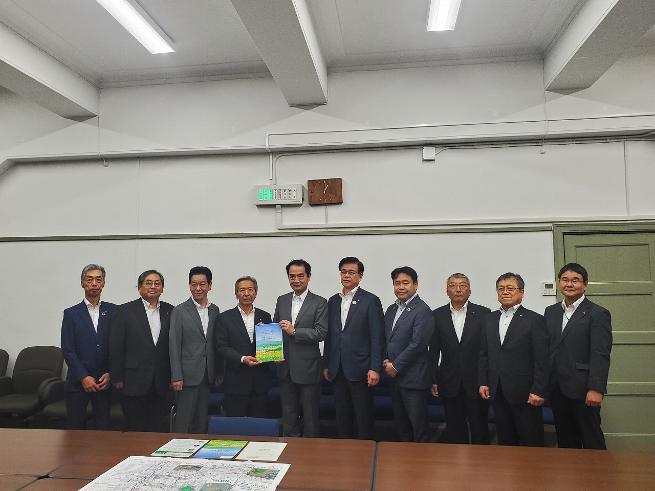 整列した男性たちが、カメラに向かって微笑みながら中央の男性が本を掲げている会議後の記念写真